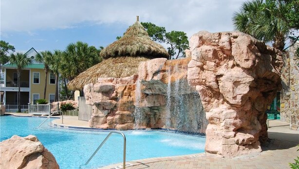 A swimming pool with a rock formation and a thatched-roof hut, surrounded by palm trees and a building with balconies. - Purple Parrot Village (Pensacola)