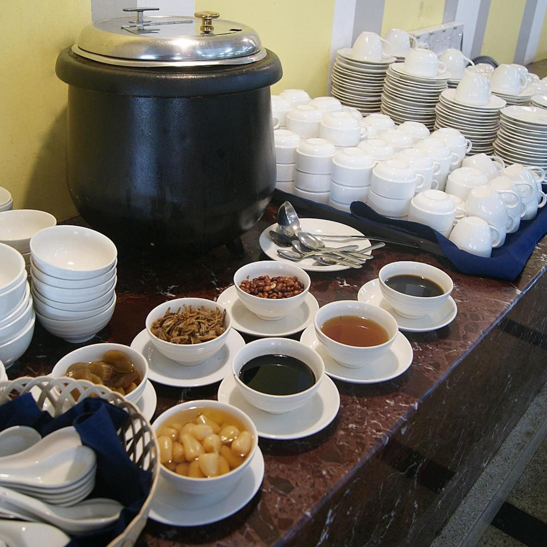 Una mesa de buffet con diversos platos, incluyendo tazones de sopa y frijoles, además de pilas de platos y tazas blancos.