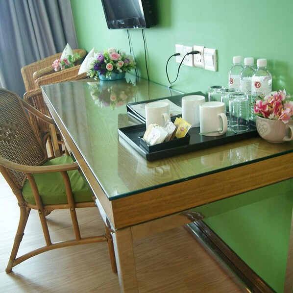 Habitación de hotel con una mesa de vidrio, sillas de mimbre, un televisor de pantalla plana en la pared y una bandeja con tazas y un arreglo floral.