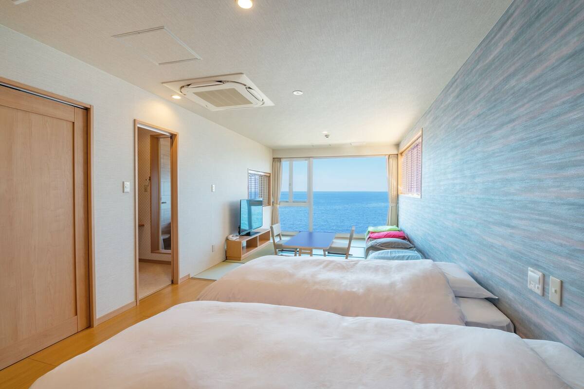 A hotel room with a large bed, a small desk with a chair, a television, and a view of the sea.