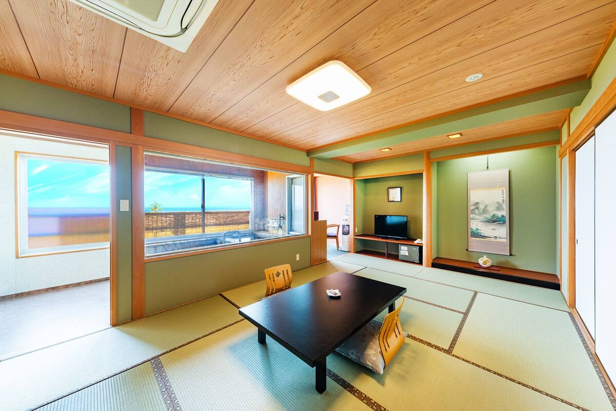 A room with tatami flooring, a low table, a television, and a large window offering a view of the sea.
