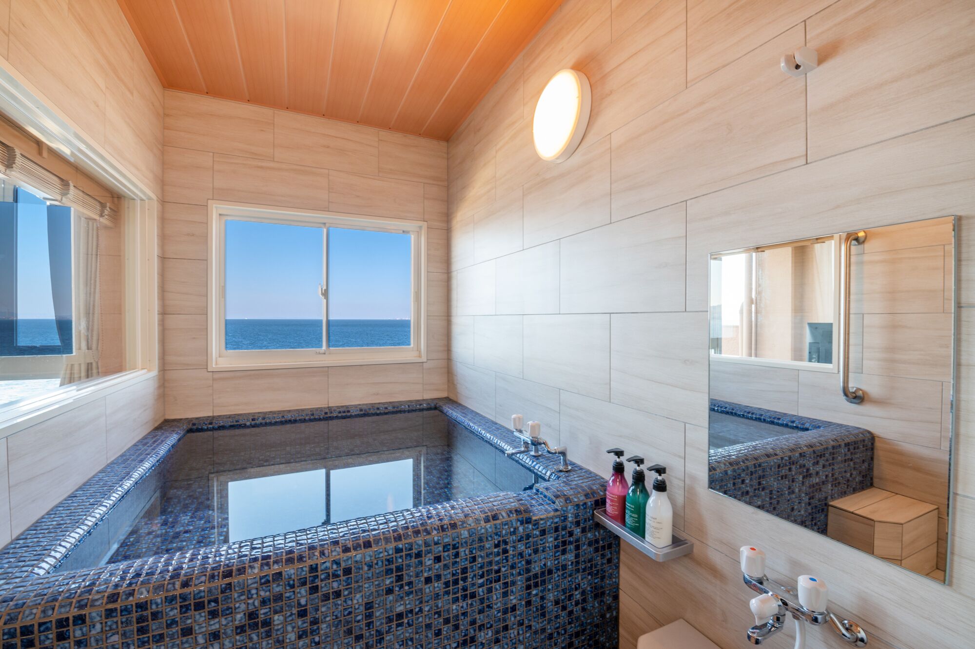 A modern bathroom with a large bathtub, a window with a view of the sea, and a mirror with a mosaic backsplash.
