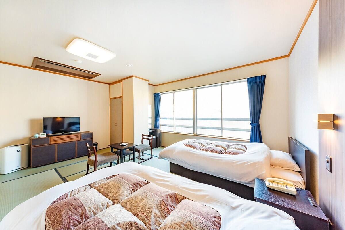 A hotel room with two beds, a desk, a television, and a large window with curtains.