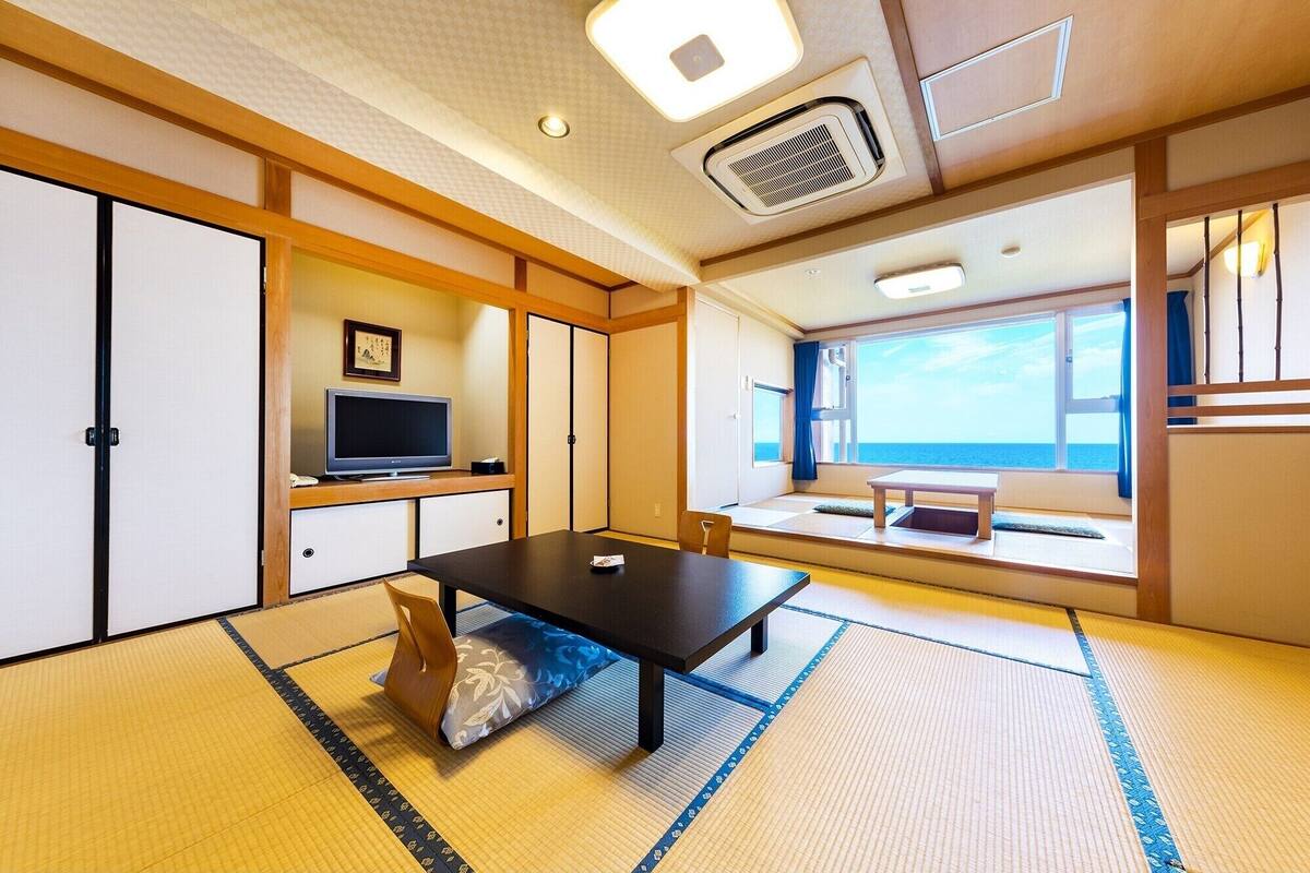 A traditional Japanese room with tatami flooring, a television, a sliding door, and a view of the sea.