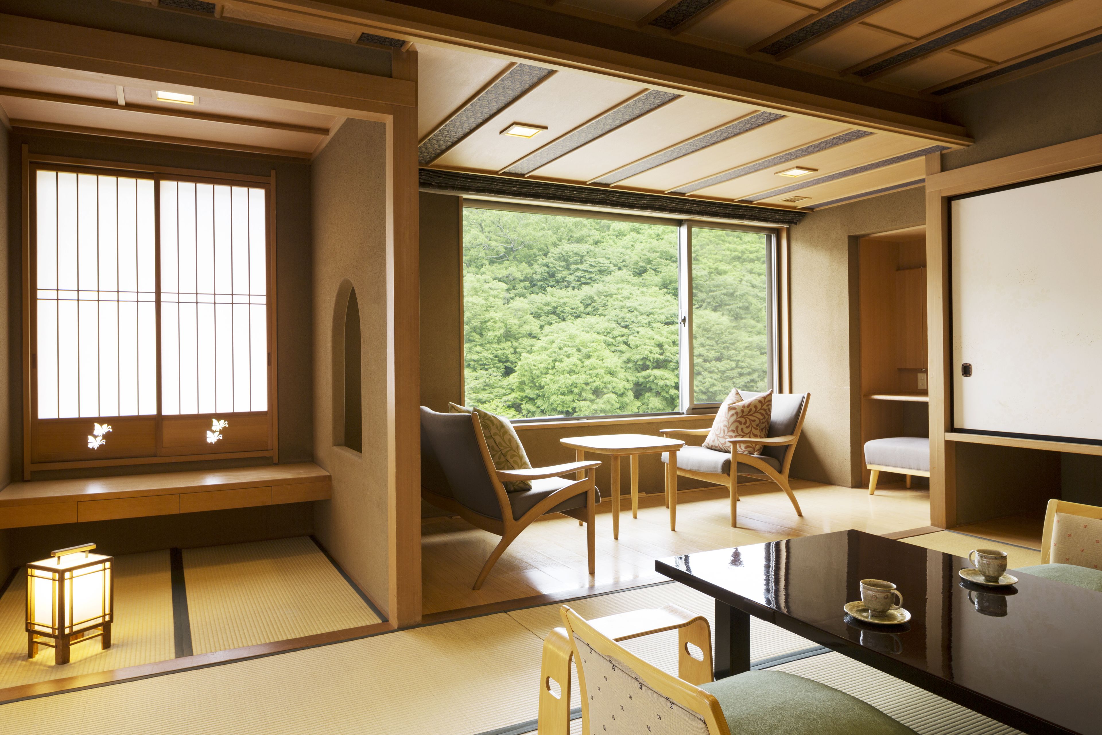 A traditional Japanese room with tatami flooring, a low table, and a view of a forest through large windows.