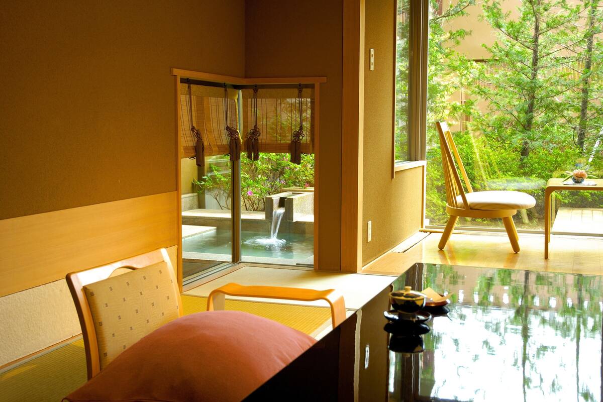 A room with a glass door leading to a garden, a table with a cushion, and a chair.