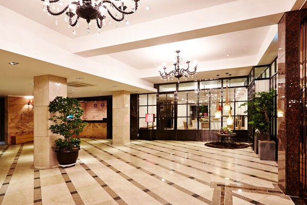 A spacious lobby with a checkered floor, large columns, potted plants, and a grand chandelier.