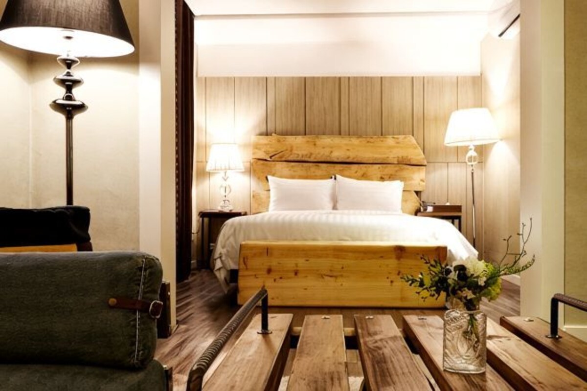 Une chambre d’hôtel avec une tête de lit en bois, un lit avec une literie blanche, un fauteuil vert, un banc en bois et un vase rempli de fleurs.