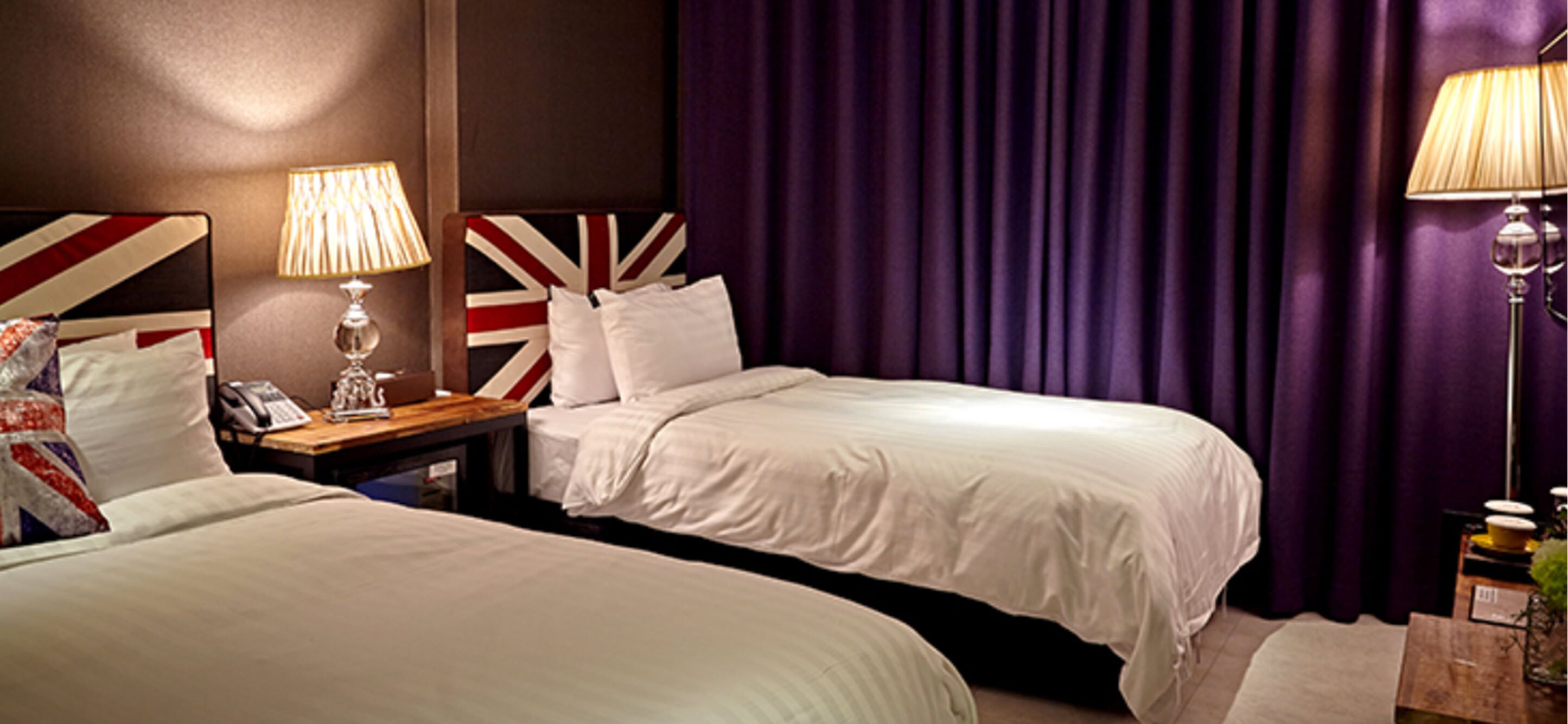 Ein Hotelzimmer mit zwei Betten, einem Nachttisch mit Lampe, einem Telefon und einem Kopfstück mit britischer Flagge.
