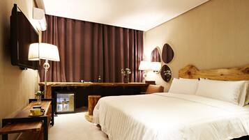 Habitación de hotel con cama, escritorio, silla, refrigerador pequeño, lámpara y espejo.