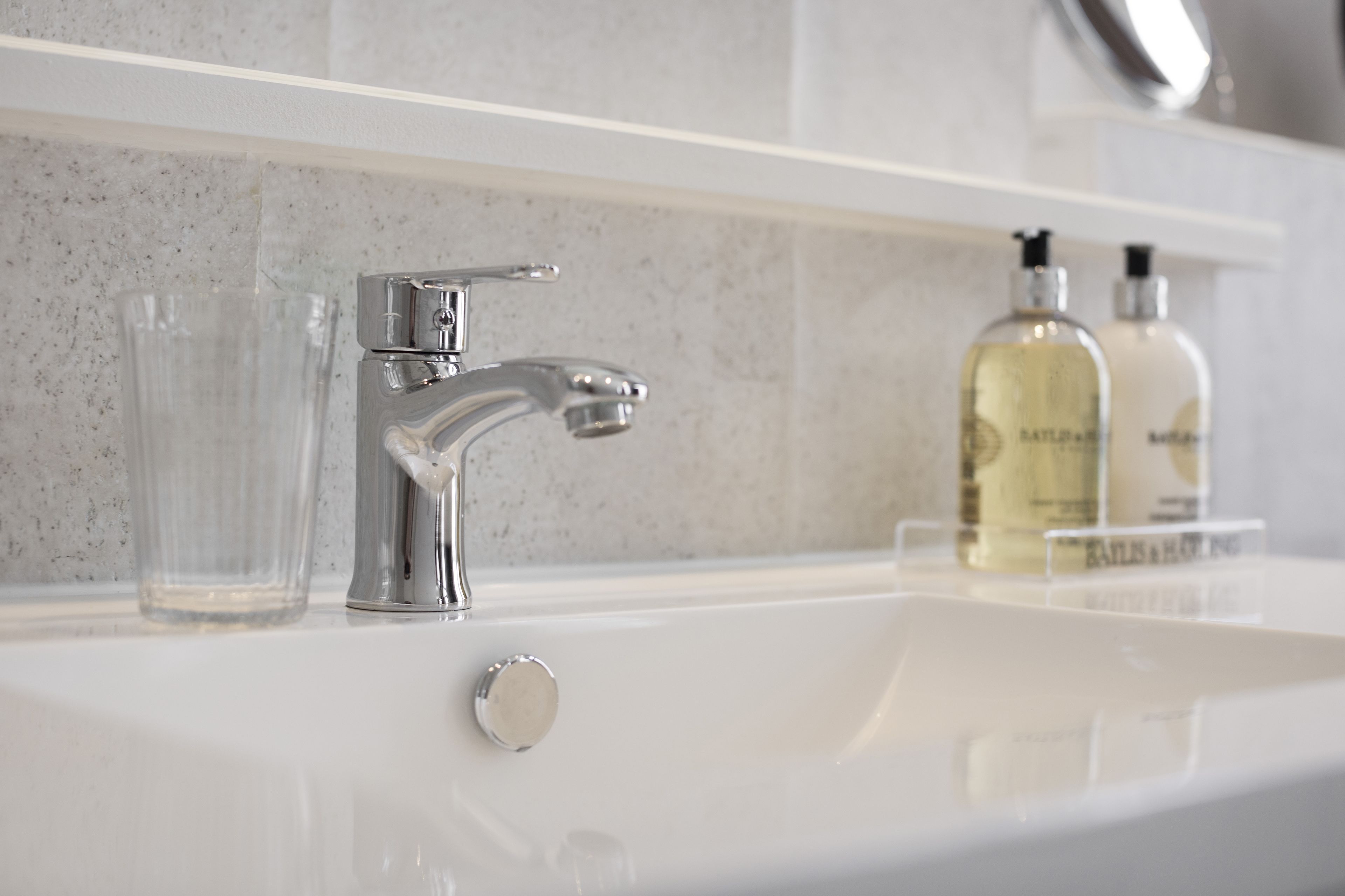 Un lavabo da bagno con rubinetto cromato, due bicchieri di vetro e due flaconi di lozione su un piano di lavoro bianco.