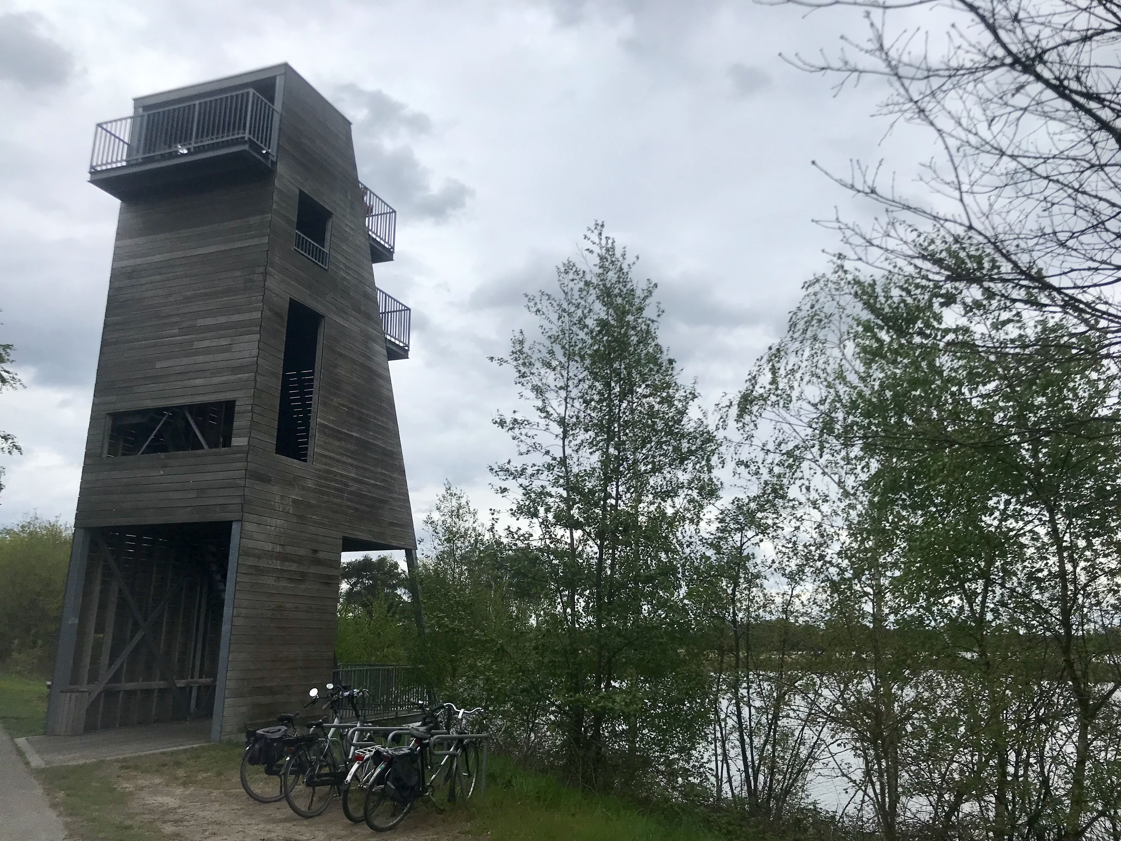 A tall, modern tower with multiple balconies and a wooden facade, surrounded by trees and a body of water.