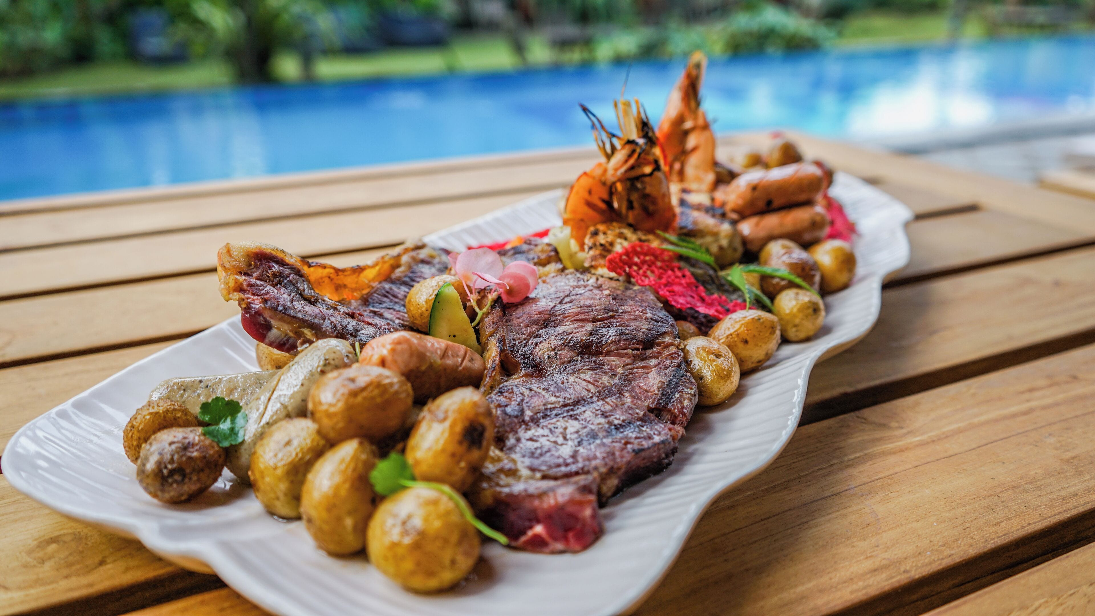 A plate of food including steak, shrimp, and potatoes, placed on a wooden table by a pool.