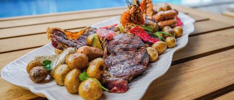 Un plato de comida con carne, camarones y papas, sobre una mesa de madera junto a una piscina.