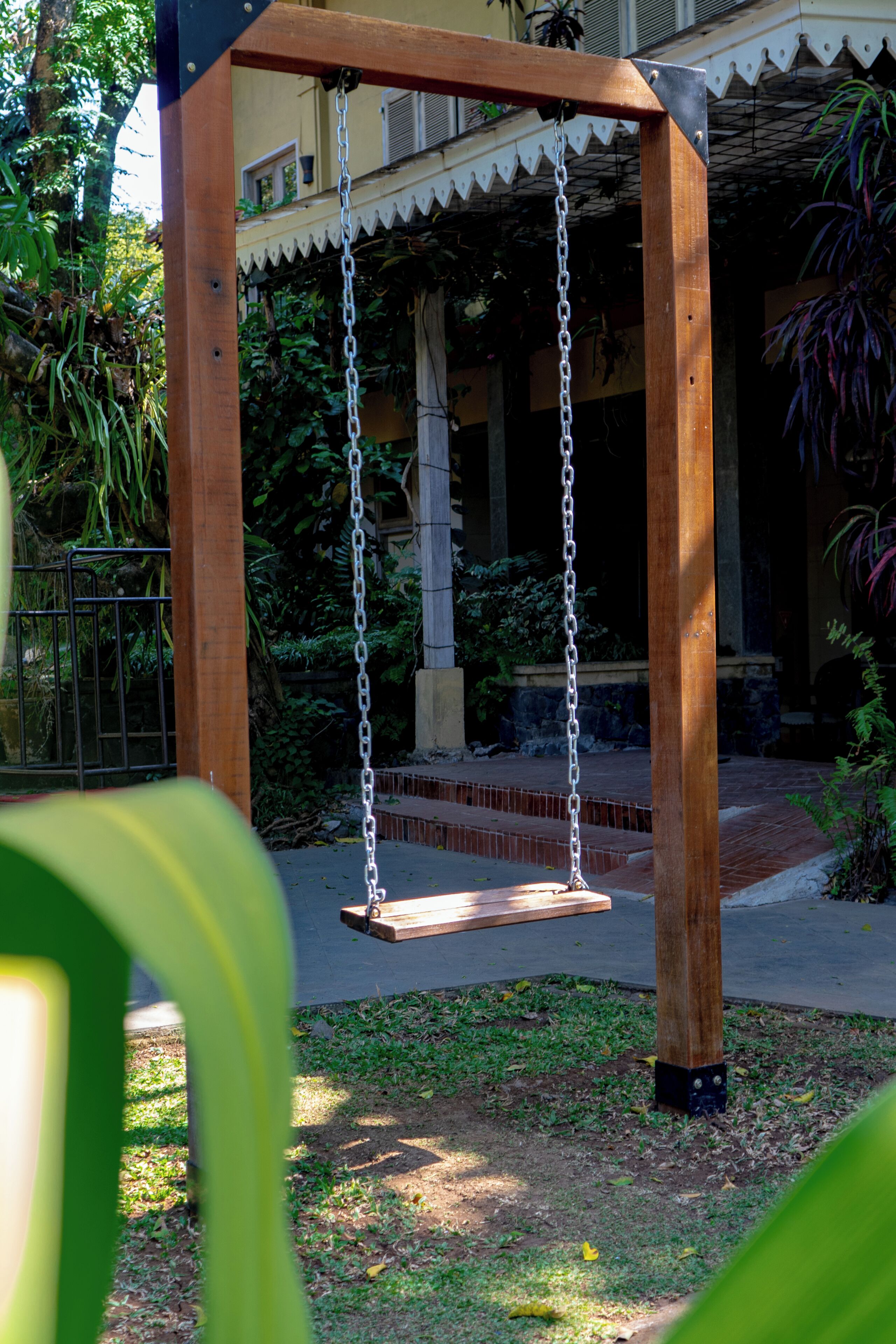 A wooden swing set with metal chains, set in a garden with a building in the background.