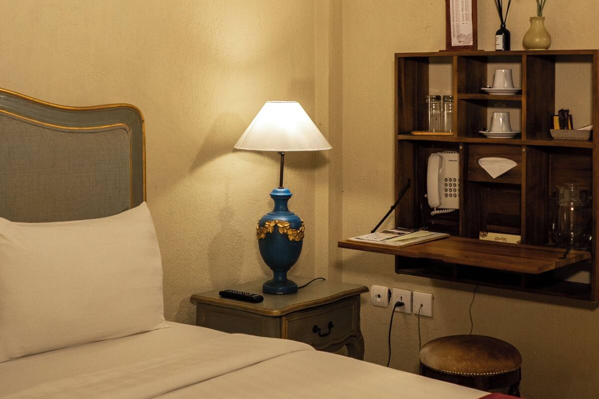 En snyggt bäddad säng med en vit kudde och ett rött överkast, en blå bordslampa, ett litet träbord, en telefon och en hylla med diverse föremål, inklusive en vas med en växt.
