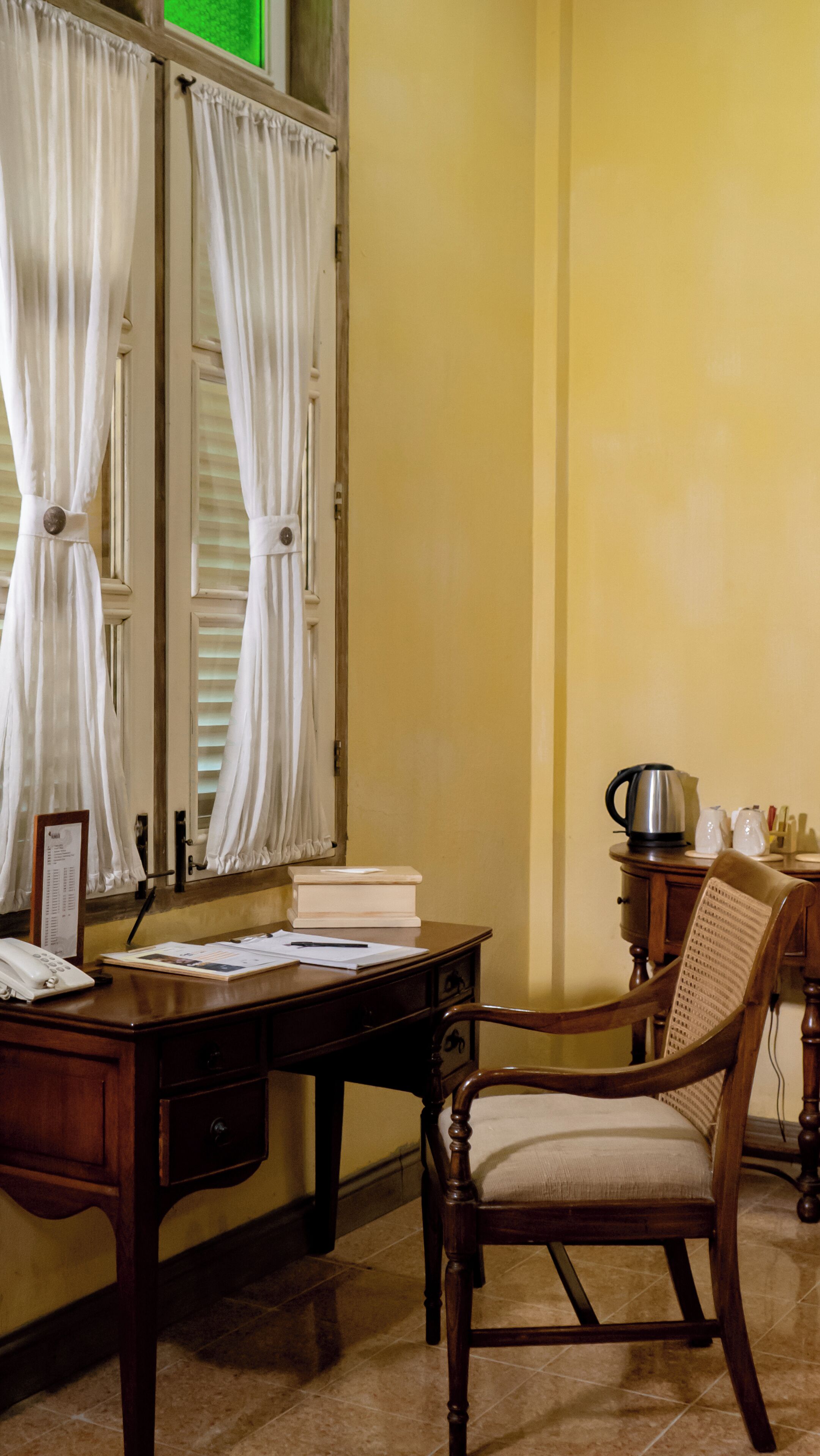 A room with a wooden desk, a chair, a telephone, and a window with white curtains.