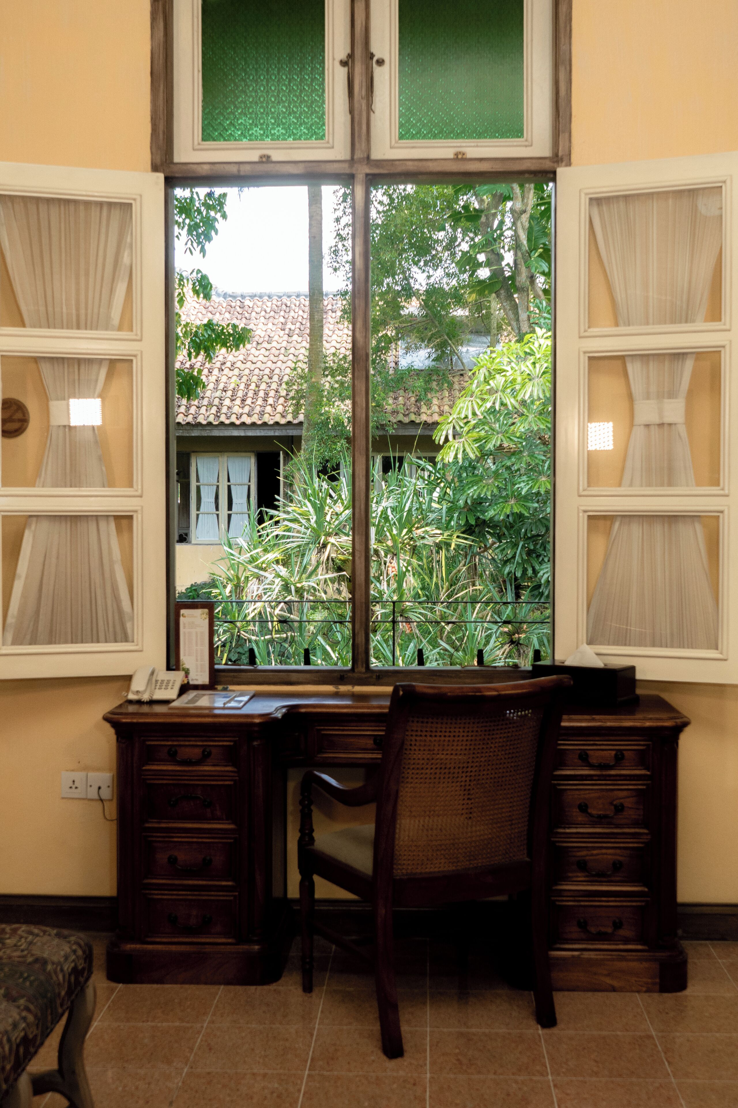 A room with a desk, chair, and a window with a view of a house and greenery.