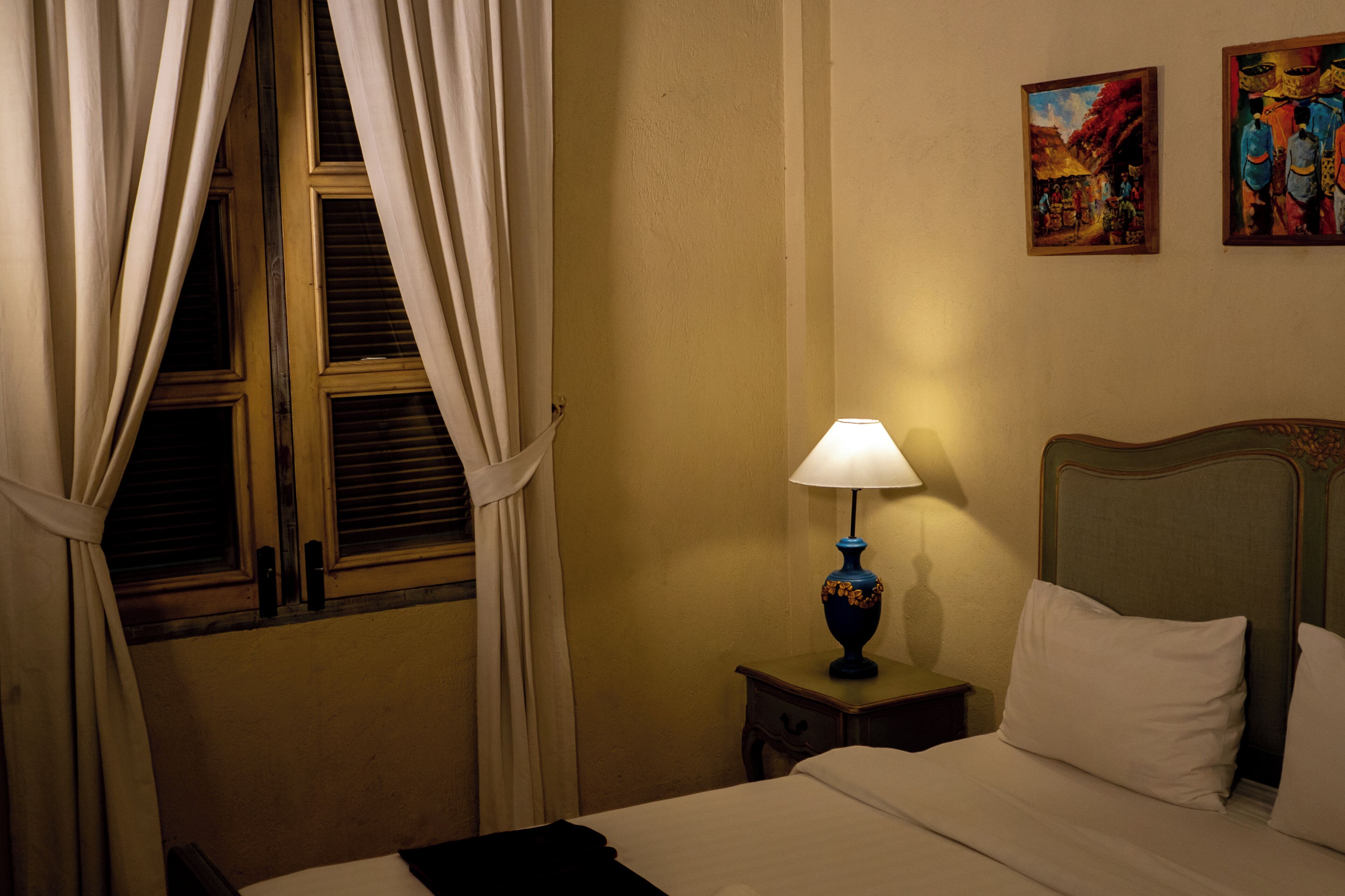 Una habitación de hotel con una cama, una mesita de noche con lámpara, una ventana con cortinas y dos cuadros en la pared.