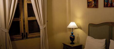 Una habitación de hotel con una cama, una mesita de noche con lámpara, una ventana con cortinas y dos cuadros en la pared.