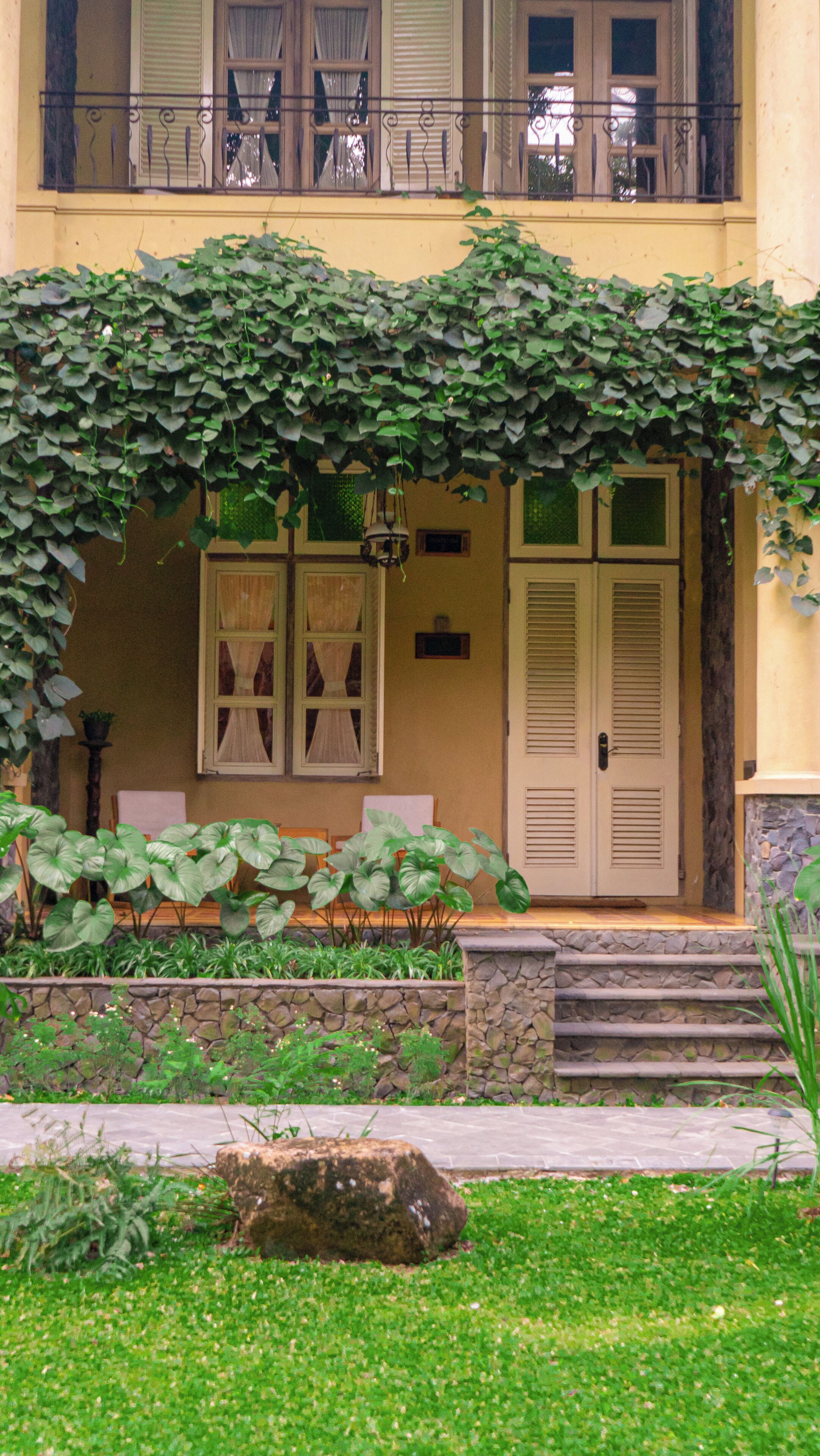 A building with a yellow facade, a balcony with metal railings, a door with shutters, and a stone wall in front.