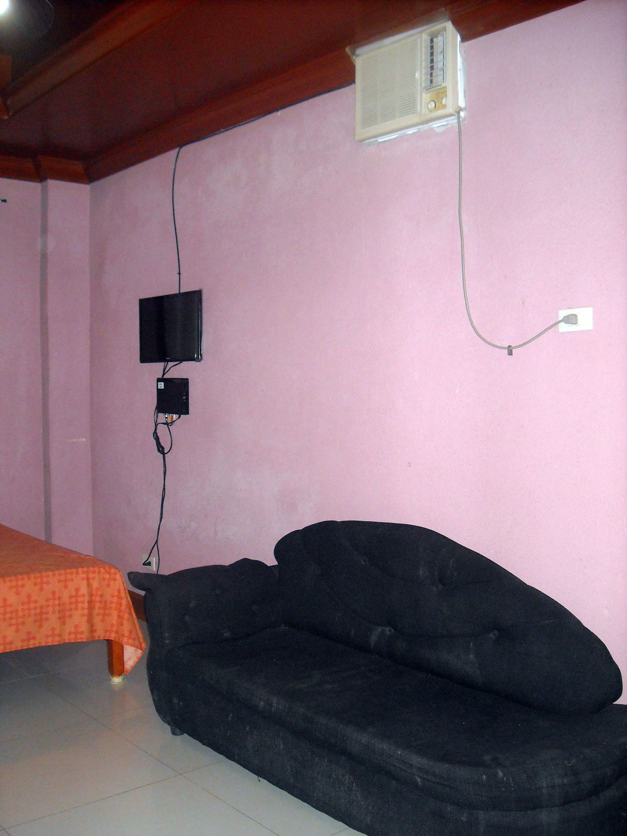 A room with a pink wall, a black sofa, a small TV mounted on the wall, and a power outlet.