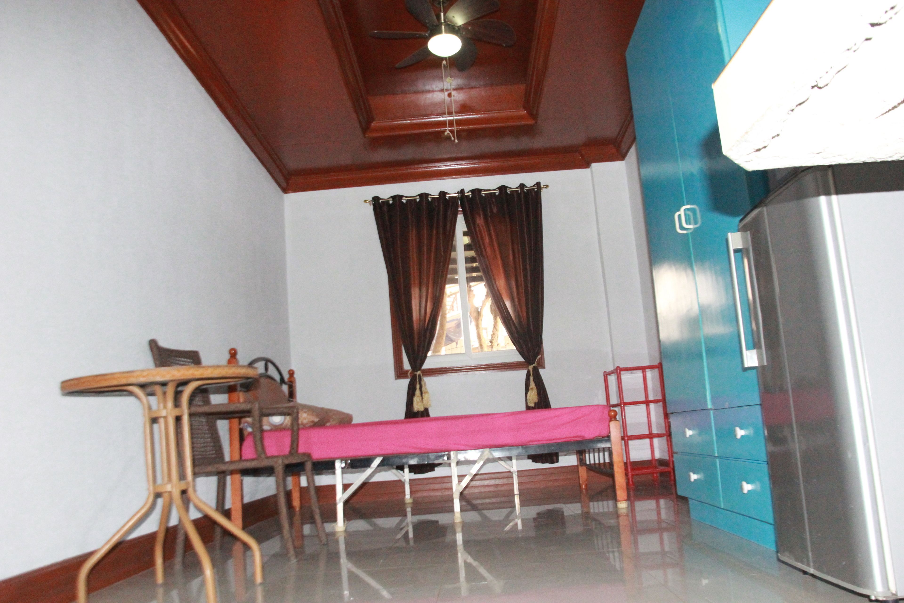 A room with a ceiling fan, a table with chairs, a bed with a pink bedspread, a window with curtains, and a refrigerator.