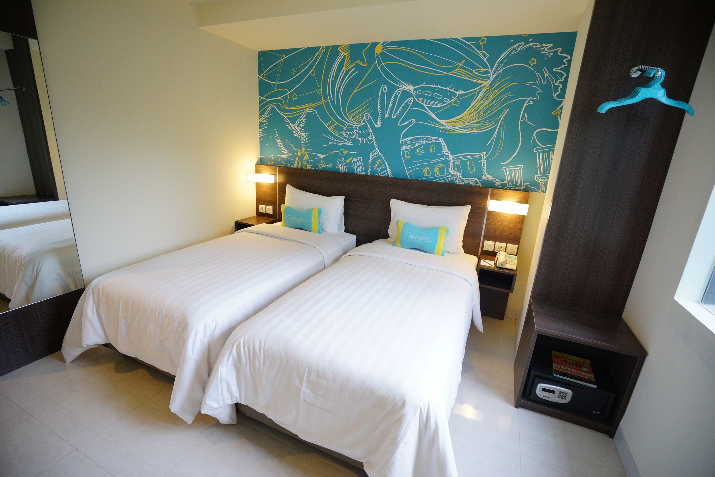 A hotel room with two beds, a large wall mural, a nightstand with a phone, and a small shelf with a book and a remote control.