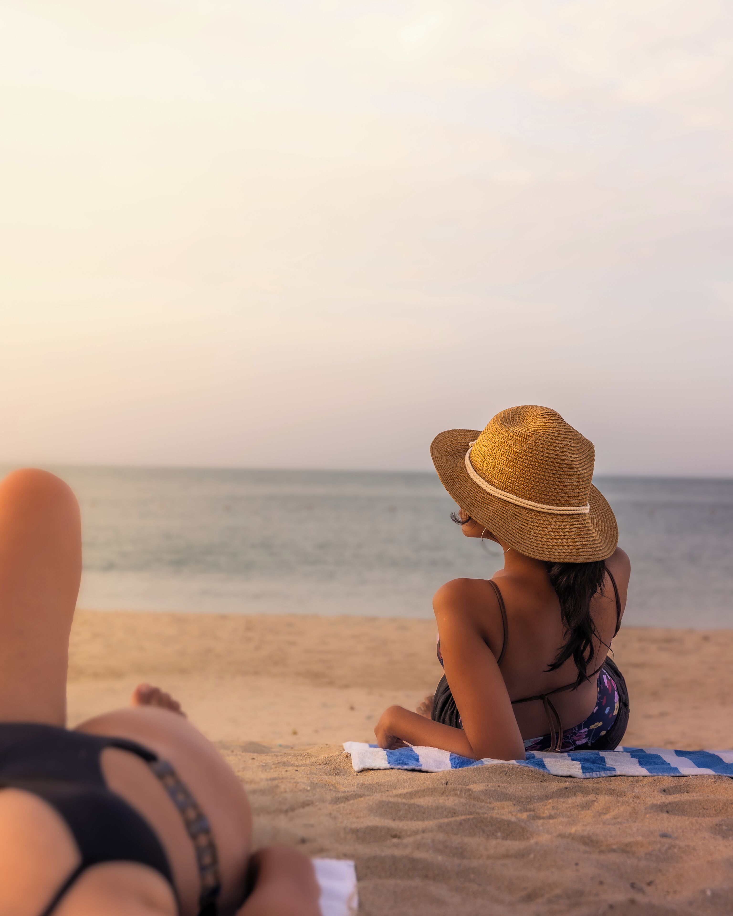 A person sitting on a beach towel with a straw hat, facing the ocean.