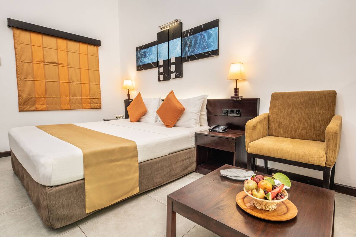 A hotel room with a bed, a chair, a nightstand, a lamp, and a fruit basket on a table.