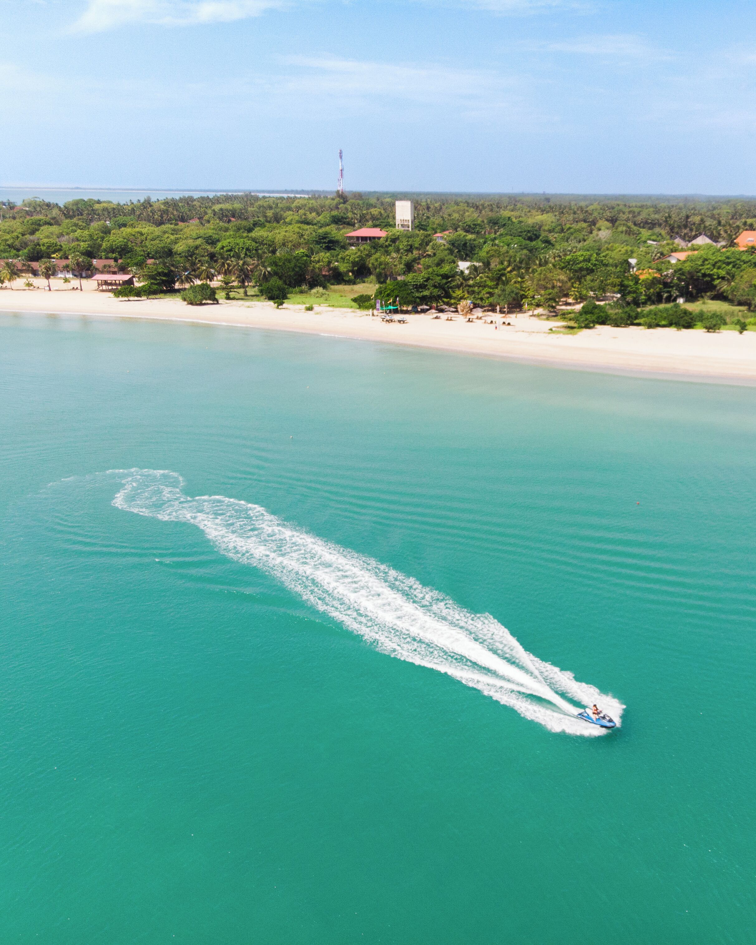 Un jet ski generando una estela en agua turquesa, con una playa y vegetación al fondo.