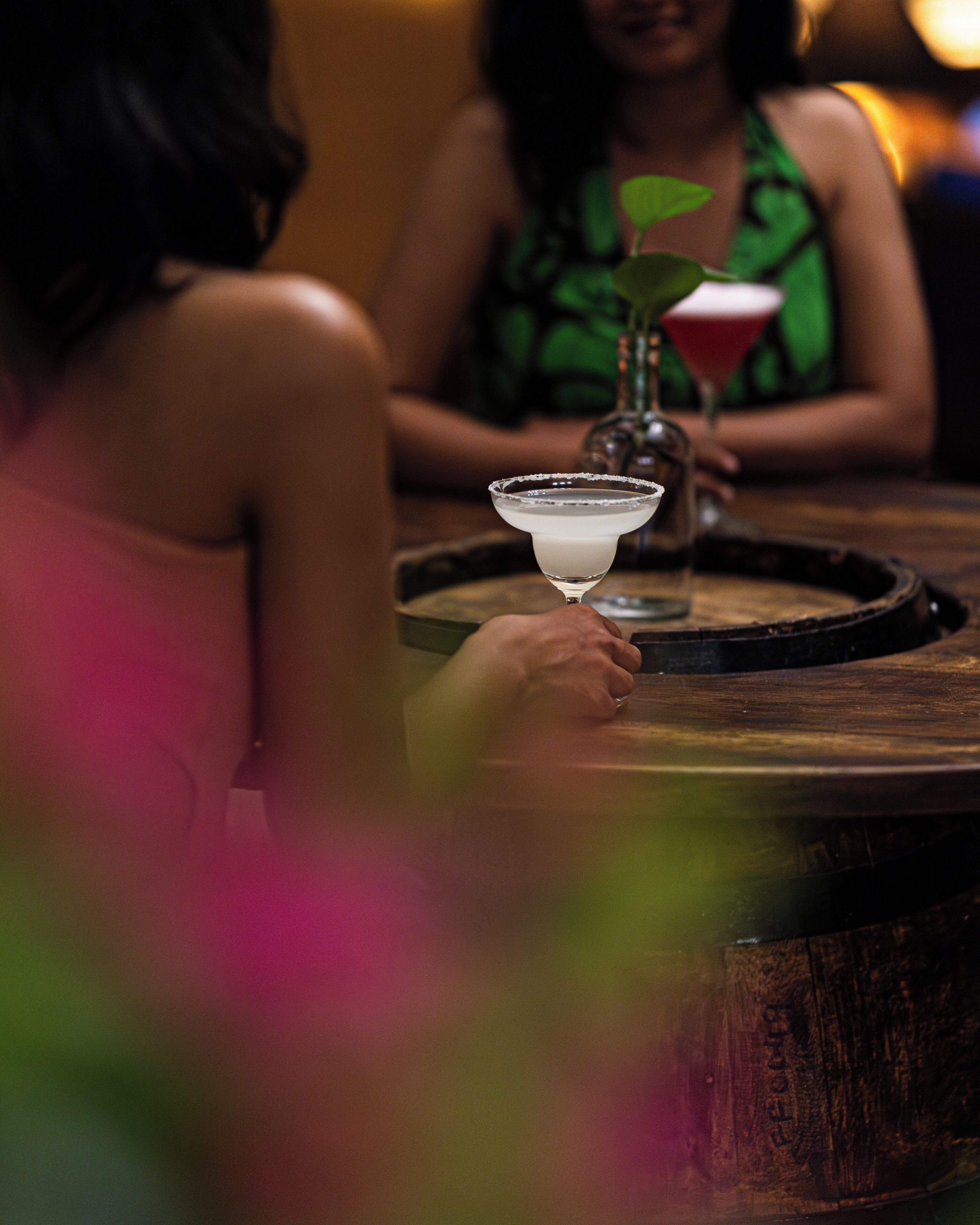 A person holding a martini glass with a salted rim, sitting at a wooden table with a bottle and a glass of red drink.