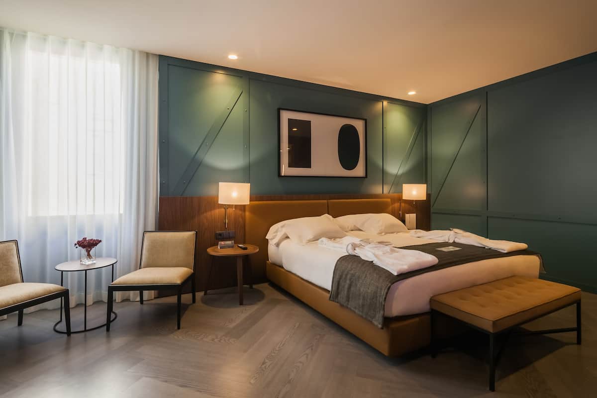Una habitación de hotel moderna con una cama grande, dos sillones, una mesita y una lámpara de noche.