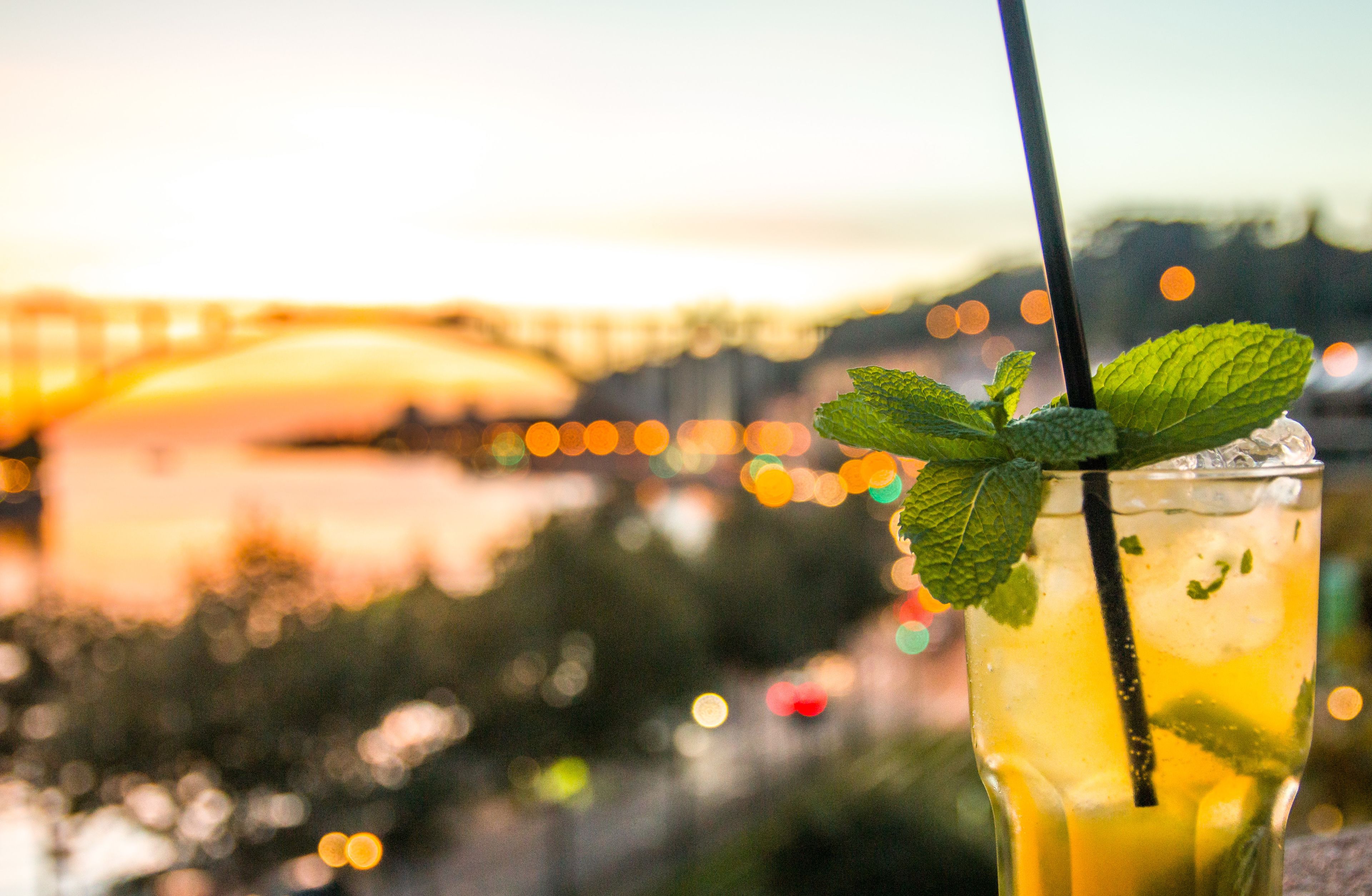 Un verre contenant une boisson jaune, orné de feuilles de menthe et muni d’une paille, le tout présenté sur un fond flou de coucher de soleil avec des lumières en arrière-plan créant un effet bokeh.