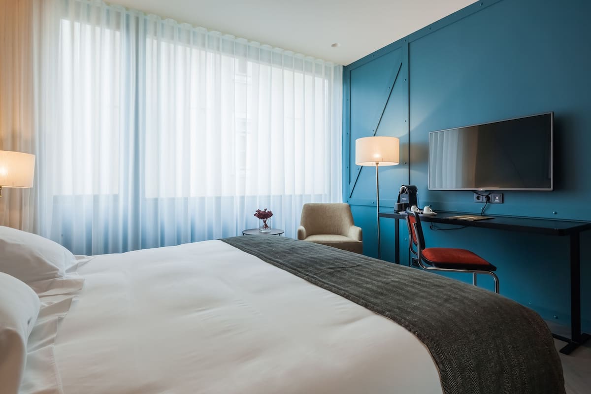 Habitación de hotel con una cama grande, un escritorio con silla, un televisor y una ventana con cortinas translúcidas.