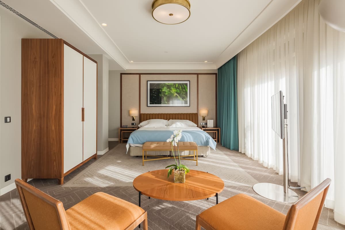 Una habitación de hotel moderna con una cama grande, un armario de madera, una mesa redonda de madera, dos sillas naranjas y una lámpara de pie.