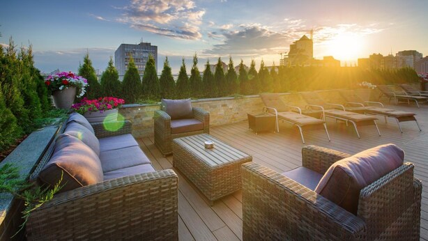 A rooftop terrace with wicker furniture, a large potted plant with flowers, and a view of the city skyline at sunset.