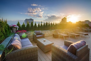 Una terraza en la azotea con mobiliario de mimbre, una planta grande con flores y vistas del horizonte urbano al atardecer.