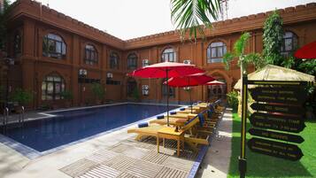 A pool area with lounge chairs and umbrellas, a sign with pool rules, and a brick building with arched windows.