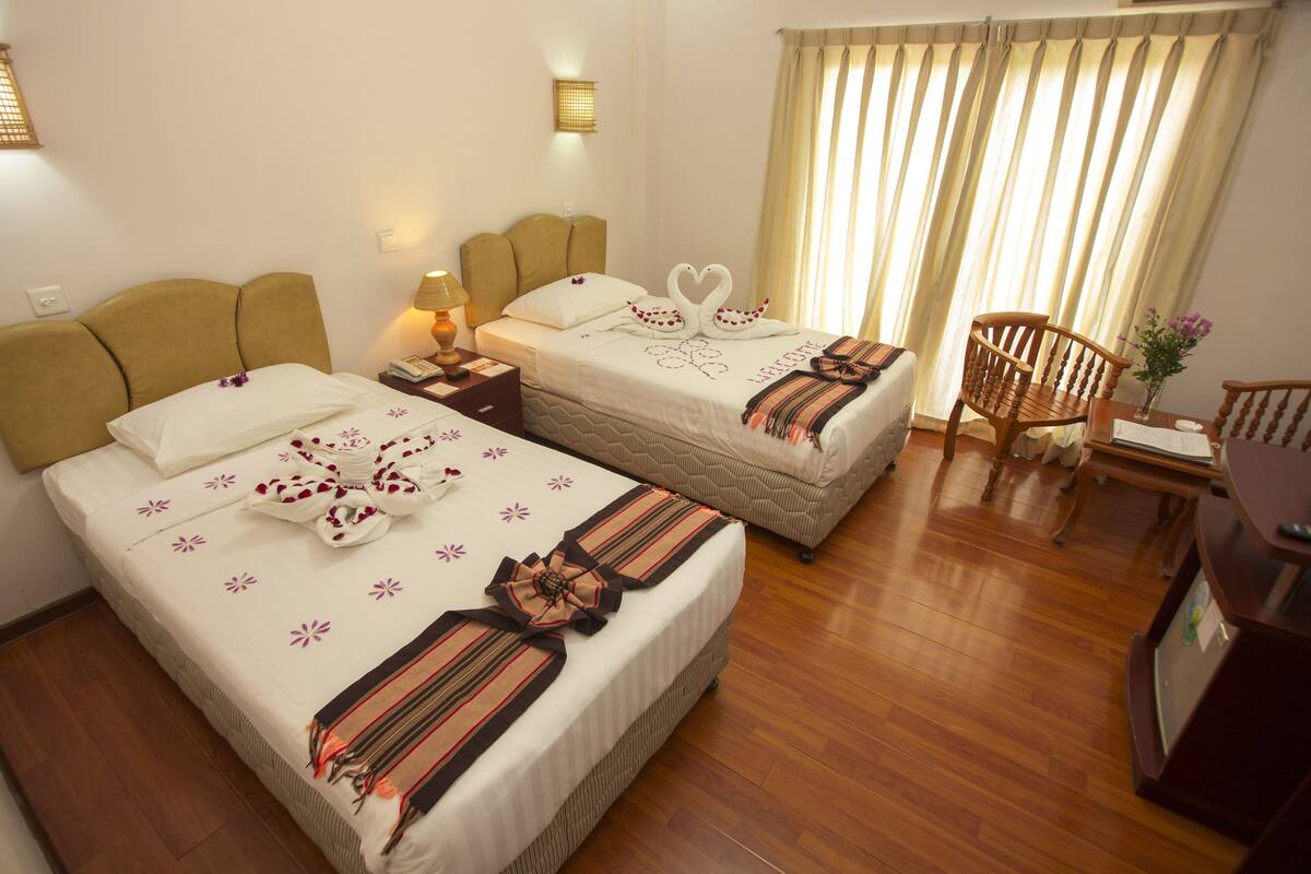 A hotel room with two beds, a wooden floor, a desk with a chair, and a window with curtains.