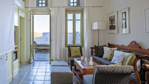 A cozy living room with a sofa, a wooden coffee table, and a dining table. There are two windows with curtains, a door leading outside, and a small balcony visible through the door. - Crossroads Inn (Tinos)