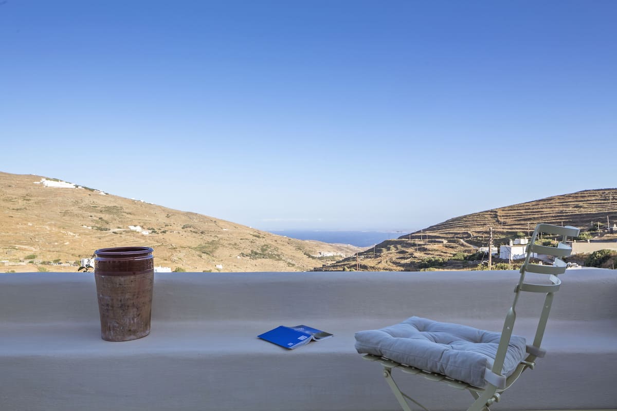Una tumbona con cojín y un libro, un jarrón grande y una vista panorámica de colinas y el mar.