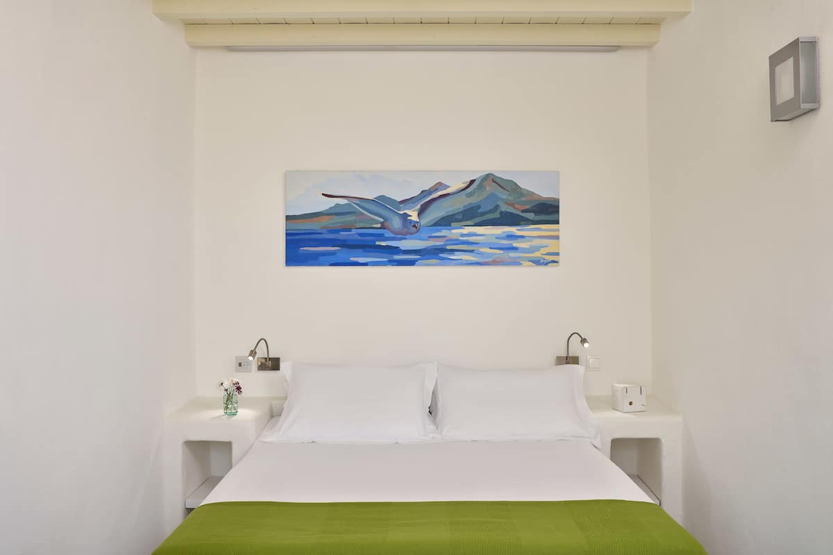 Un dormitorio con cama, mesitas de noche blancas, un cuadro de montañas y un pájaro, y una colcha verde.