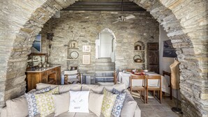 A stone-walled room with a sofa, dining table, and a ceiling fan. - Crossroads Inn (Tinos)