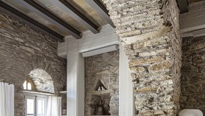 A rustic interior with exposed stone walls, a wooden ceiling, a sofa with patterned cushions, and a dining table with chairs. - Crossroads Inn (Tinos)