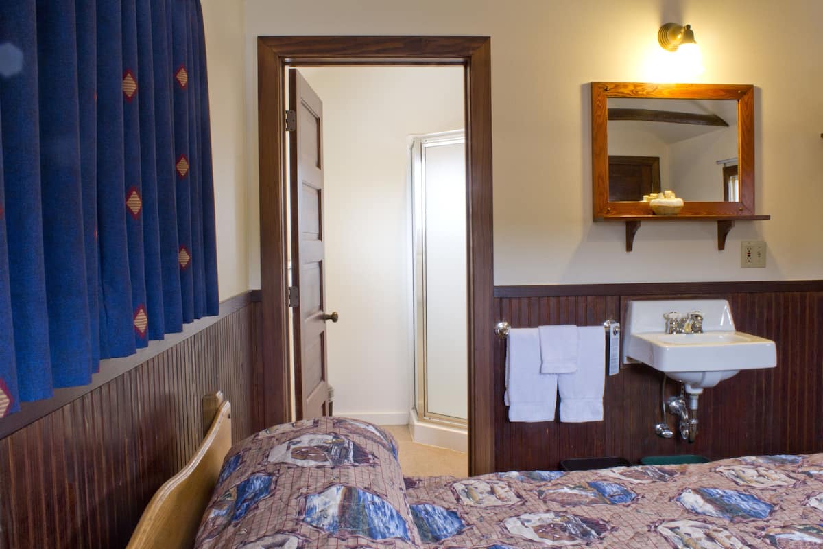 Una habitación de hotel con una cama, cabecero de madera, baño con lavamanos y espejo, y una puerta que da a otra habitación.