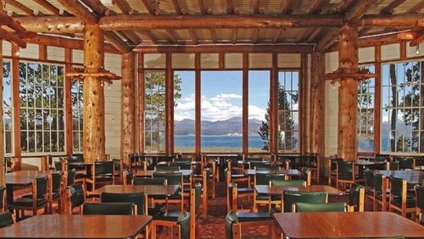 Un restaurante con mesas y sillas de madera, grandes ventanales con vistas a montañas y un cuerpo de agua.