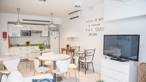 Una cucina moderna con mobili bianchi, un forno a microonde e un condizionatore d'aria. C'è una zona pranzo con sedie e tavolo bianchi, e un televisore su un mobiletto bianco.