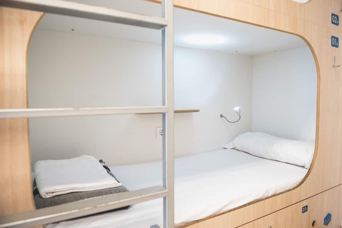 A bunk bed with a white mattress and pillow, a small shelf with folded clothes, and a wall-mounted lamp.