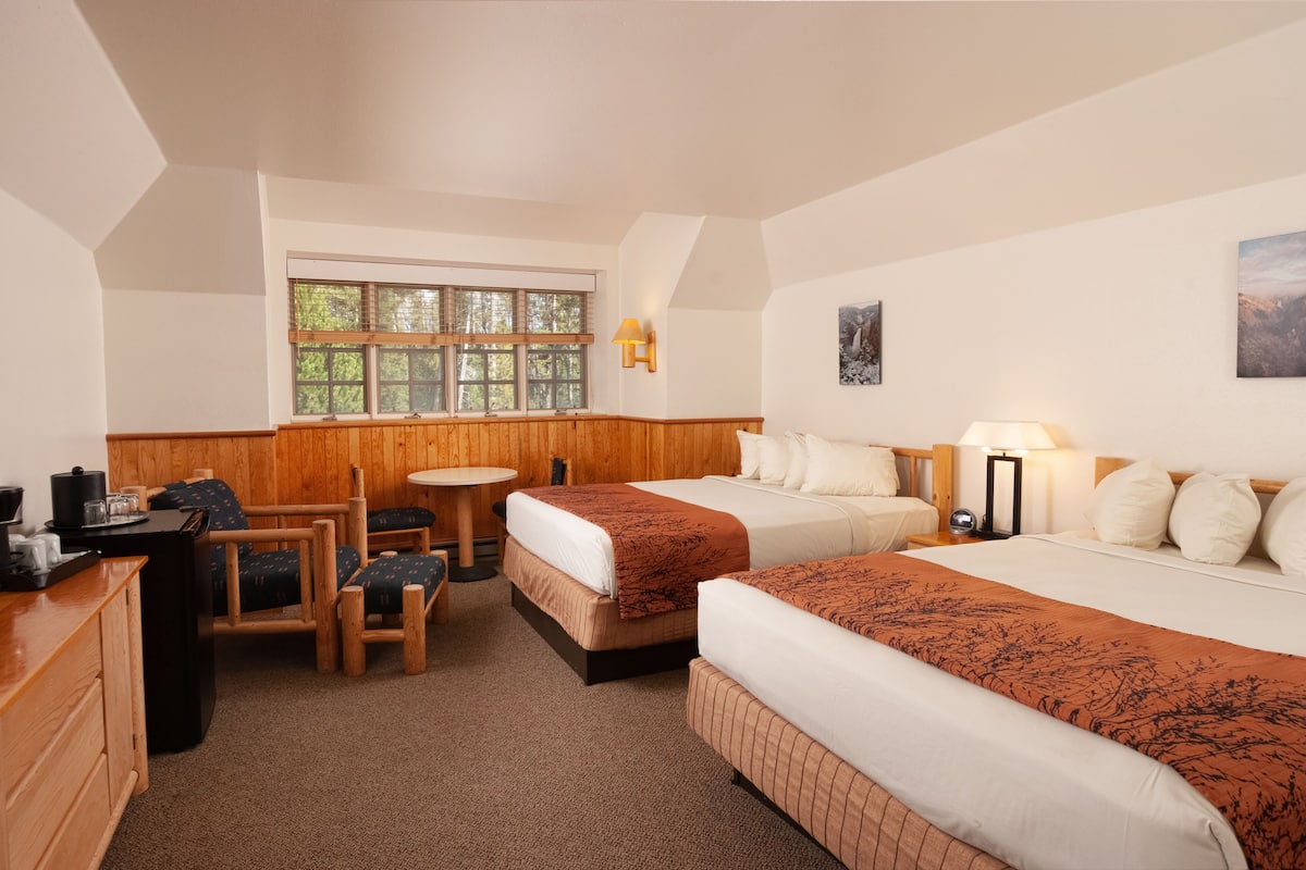 Ett hotellrum med två sängar, ett skrivbord, en stol, ett litet bord och ett fönster med utsikt över träd.